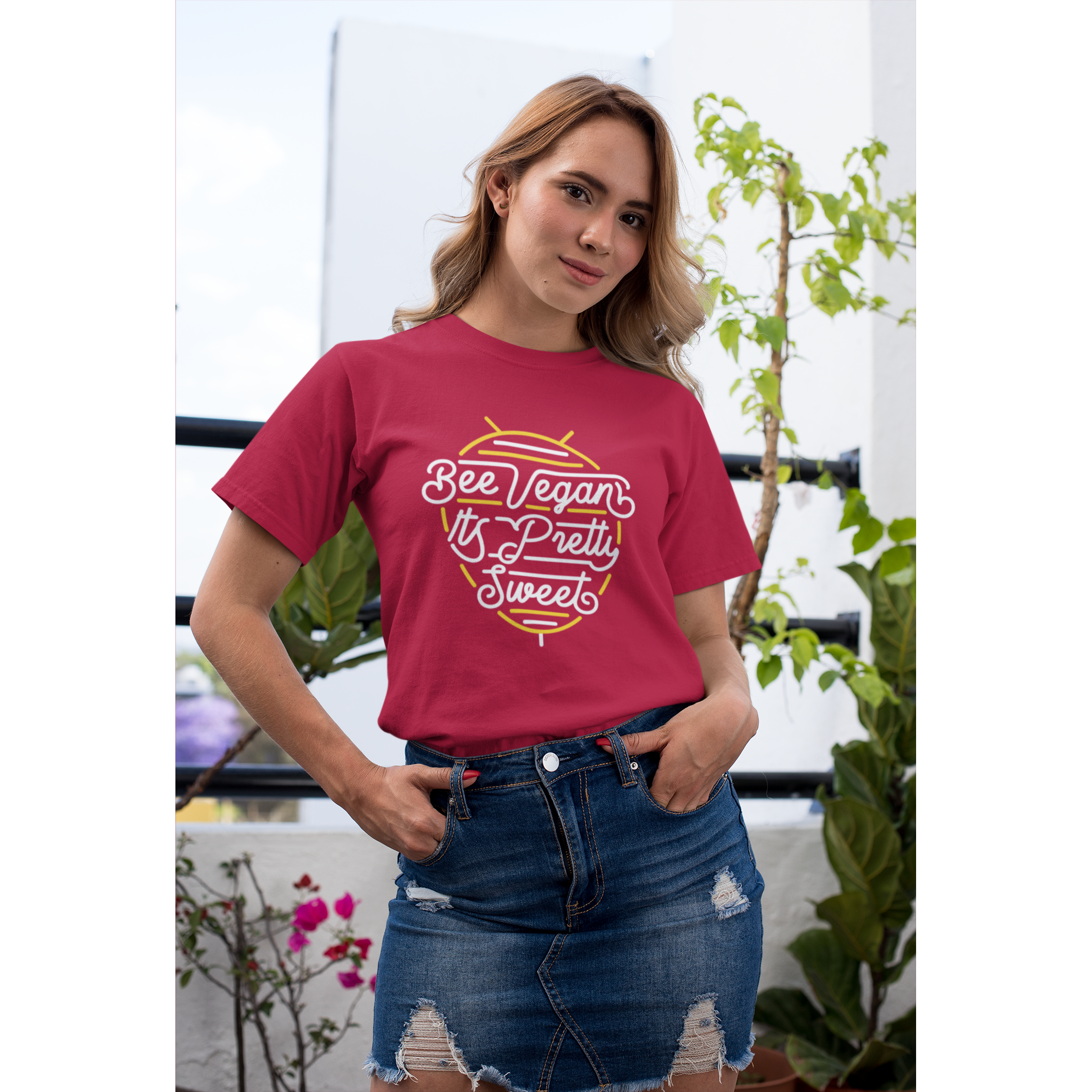 cool young tan woman standing on balcony with plants wearing bee vegan it's pretty sweet neon sign graphic design on red colored vegan t shirt from vegan shirts from ethical clothing brands