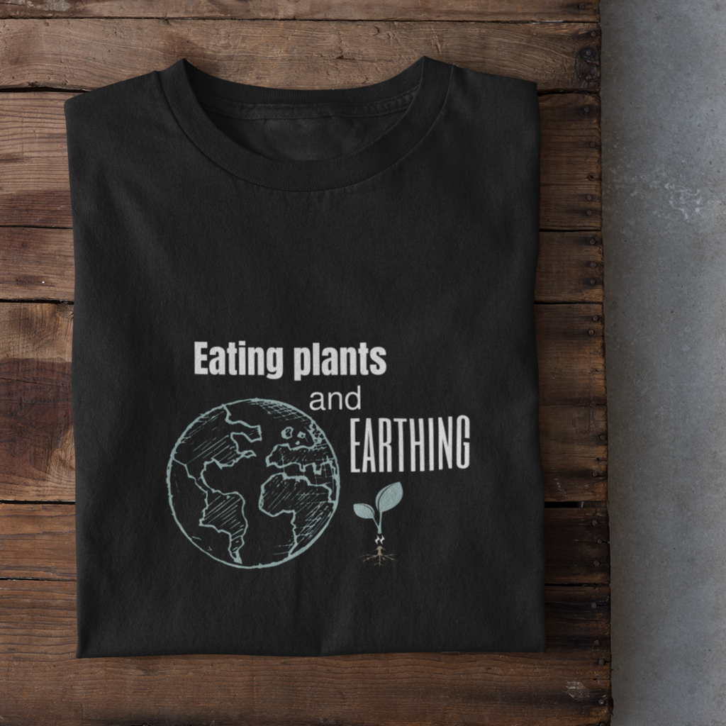 vegan shirts design saying eating plants and earthing in white lettering with mint colored earth and plant icons, on a black premium tee, folded on a wooden table with shoes