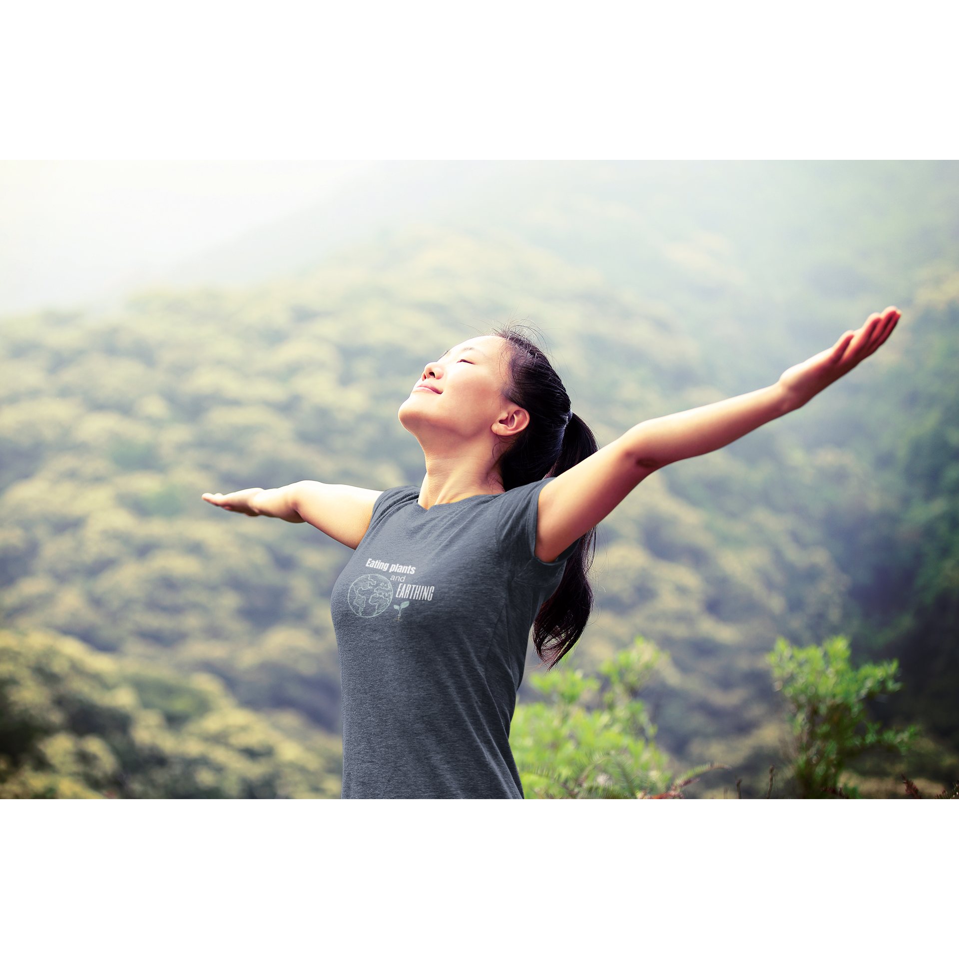 young Asian woman in woods stretching her arms out to the sky wearing an eating plants and earthing vegan t shirt design on heather navy color premium cotton t shirt for those who eat plants and enjoy their earthing mat