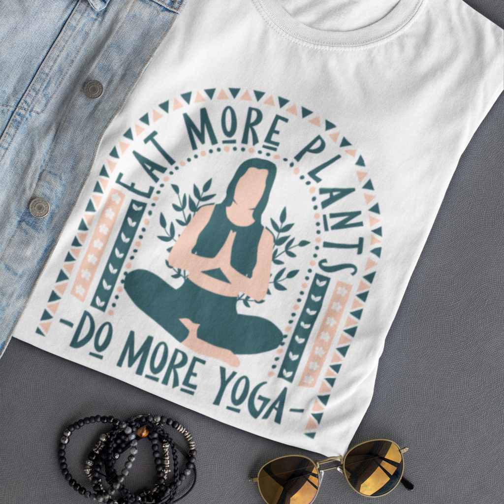 Dark grey and light muted pink boho tops design saying eat more plants, do more yoga, on a white vegan t shirt, folded, laying on a grey surface with accessories, showing vegan t shirt from vegan clothing brands