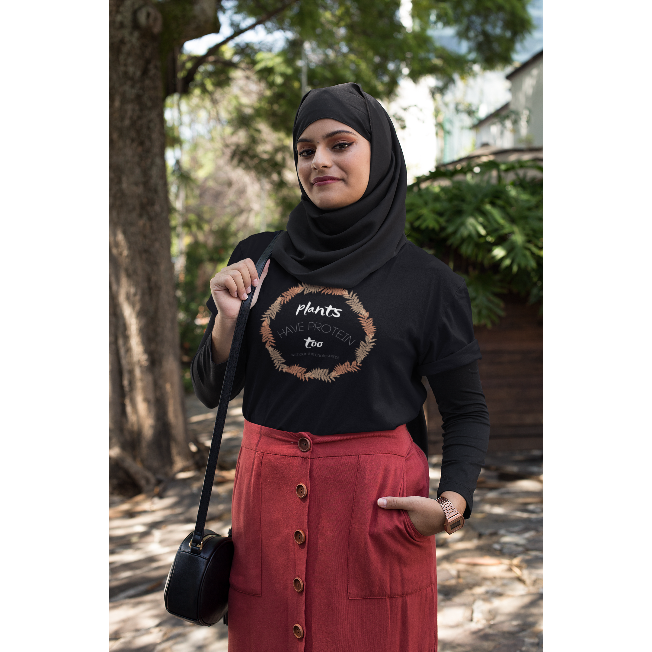 woman wearing hijab in a park, wearing a black colored vegan t shirt with white lettering and tan and peach fern circle, saying plants have protein too without the cholesterol