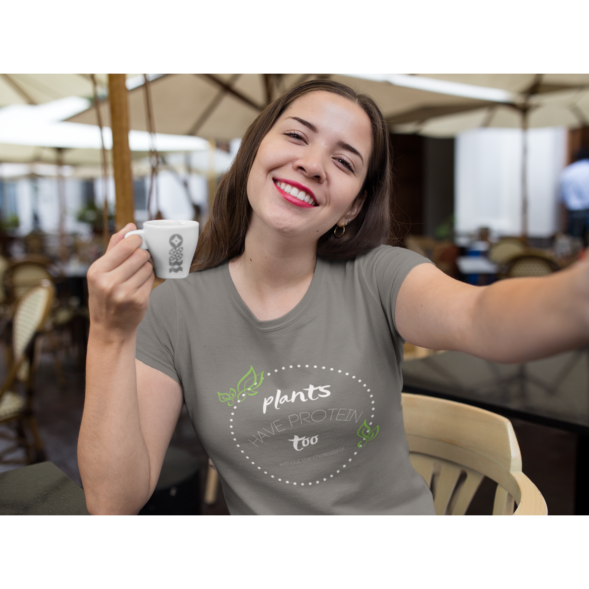 Hispanic woman drinking expresso at outdoor cafe, wearing asphalt colored vegan shirt with white lettering and polka dots, and green leaves, saying plants have protein too without the cholesterol