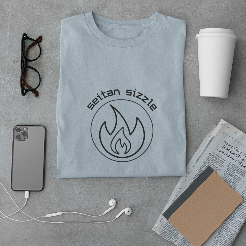 for vegans who love seitan recipes, black lettering saying seitan sizzle over a circle with flame design for vegan shirts on a folded light blue premium tee with office accessories on a cement surface